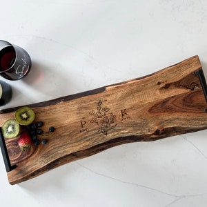 May include: A wooden serving board with a live edge, featuring a floral design and the initials "P K" with "EST. 2019" engraved. The board has black metal handles and is accompanied by fruit and a glass of red wine.