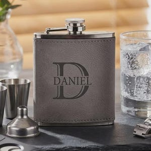 May include: A grey leather flask engraved with the name "DANIEL" and a large letter "D". The flask is accompanied by two small stainless steel shot glasses, a glass of water with ice and a lime wedge, a cigar cutter, and a watch.