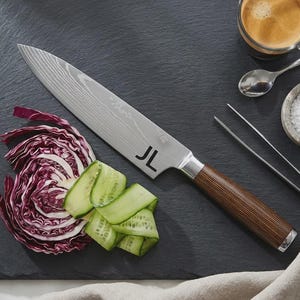Peut inclure: Un couteau de chef en acier inoxydable avec une lame à motifs et un manche en bois, à côté de tranches de concombre et de chou rouge sur une planche à découper noire. Les initiales "JL" sont gravées sur la lame. Une tasse de café, du sel et une pince sont également présents.