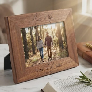 Peut inclure: Un cadre photo en bois avec une photo d'un couple se promenant dans une forêt. Le cadre porte les noms "Amelia & Kyle" et la citation "Love never fails." de 1 Corinthiens 8:8.