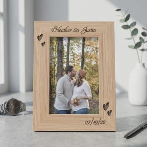 May include: A light-coloured wooden picture frame featuring the engraved text Heather & Justin at the top and 07/19/22 at the bottom. The frame includes laser-cut heart shapes on the top left and bottom right corners, holding a photograph of a couple.
