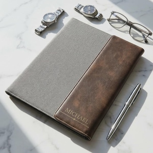 May include: A rectangular portfolio case featuring a gray fabric section and a dark brown leather-like section with the name MICHAEL engraved on the front. The case is accompanied by two silver wristwatches, clear-framed eyeglasses, and a silver pen.