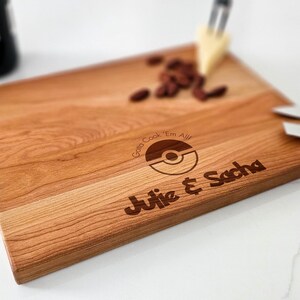 Close-up of the laser engraving on a cutting board showing the playful "Gotta Cook 'Em All!" message with couple names and date in a Pokémon style.