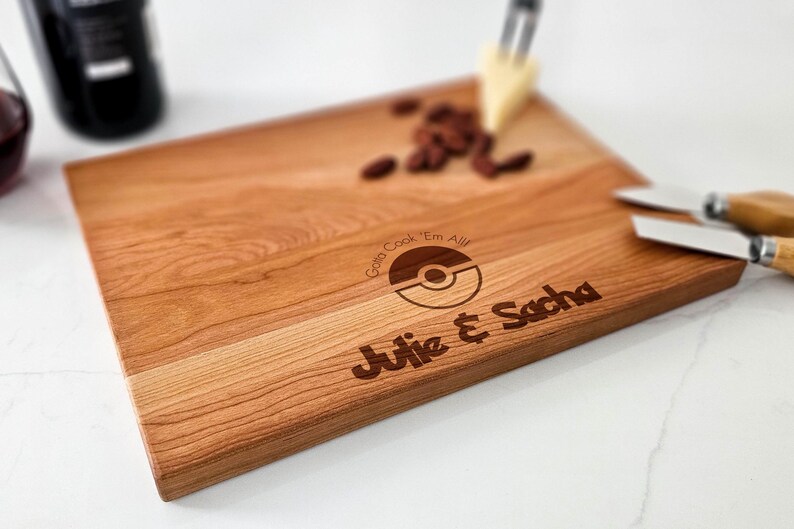 Close-up of the laser engraving on a cutting board showing the playful "Gotta Cook 'Em All!" message with couple names and date in a Pokémon style.
