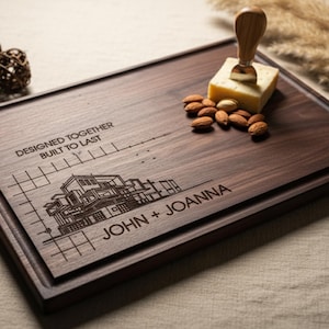 May include: A rectangular wooden cutting board with a dark brown finish. The board features engraved text and a house design, with the names "JOHN + JOANNA" below. A cheese knife and cheese are on the board, along with a scattering of almonds.
