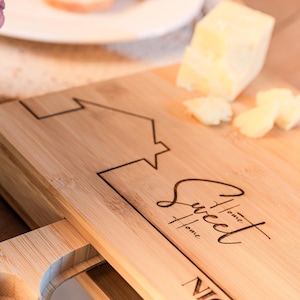 May include: A wooden chopping board with a drawer for storing knives. The board is engraved with the text "One Sweet Home" and a house design. The board also has the name "Nicole &" engraved on it.