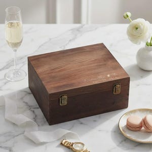 May include: A dark brown wooden box with a rectangular shape, secured by two brass-colored clasps. The box has a natural wood grain finish and features engraved text: "Samantha & Max" and "April 29 2024". The box is on a marble surface.