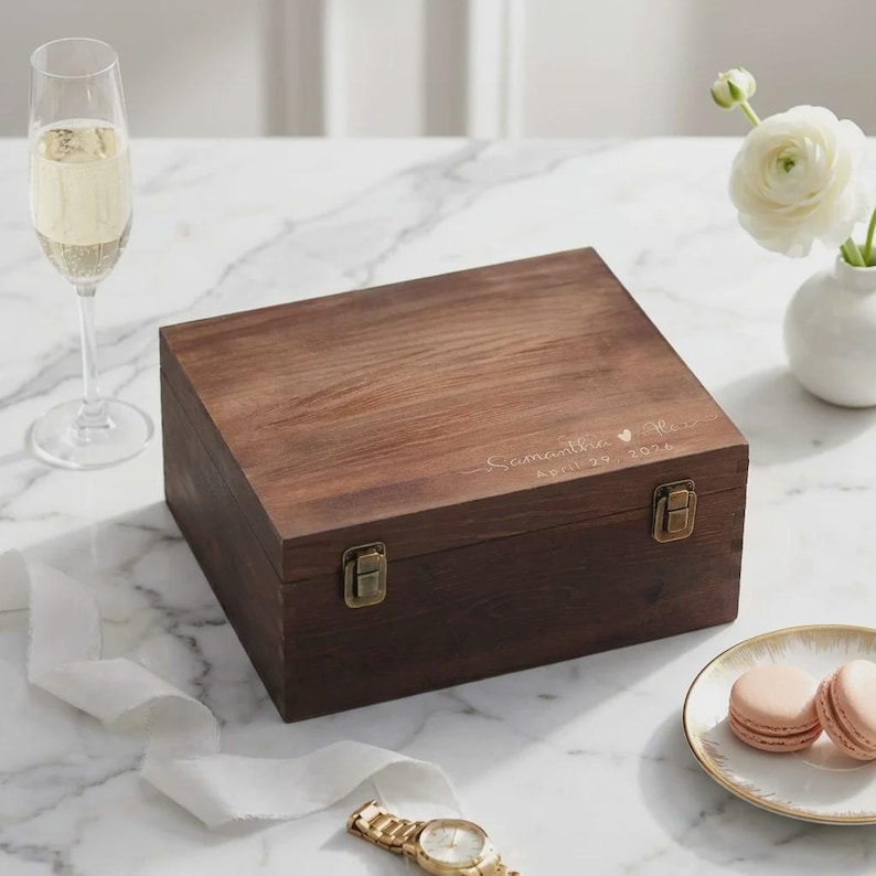 May include: A dark brown wooden box with a rectangular shape, secured by two brass-colored clasps. The box has a natural wood grain finish and features engraved text: "Samantha & Max" and "April 29 2024". The box is on a marble surface.