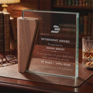 Peut inclure: Un prix de retraite en verre et en bois. Le prix comporte une base en bois et un panneau en verre transparent avec le texte "RETIREMENT AWARD" et "ADAM BRENT". Le prix est sur une surface brune.