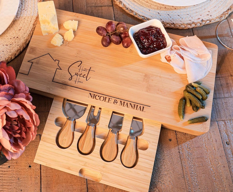 May include: A wooden cheese board with a built-in drawer for cheese knives. The board has the text "Home Sweet Home" and "Nicole & Manhal" engraved on it. The board is filled with cheese, grapes, crackers, and other snacks.