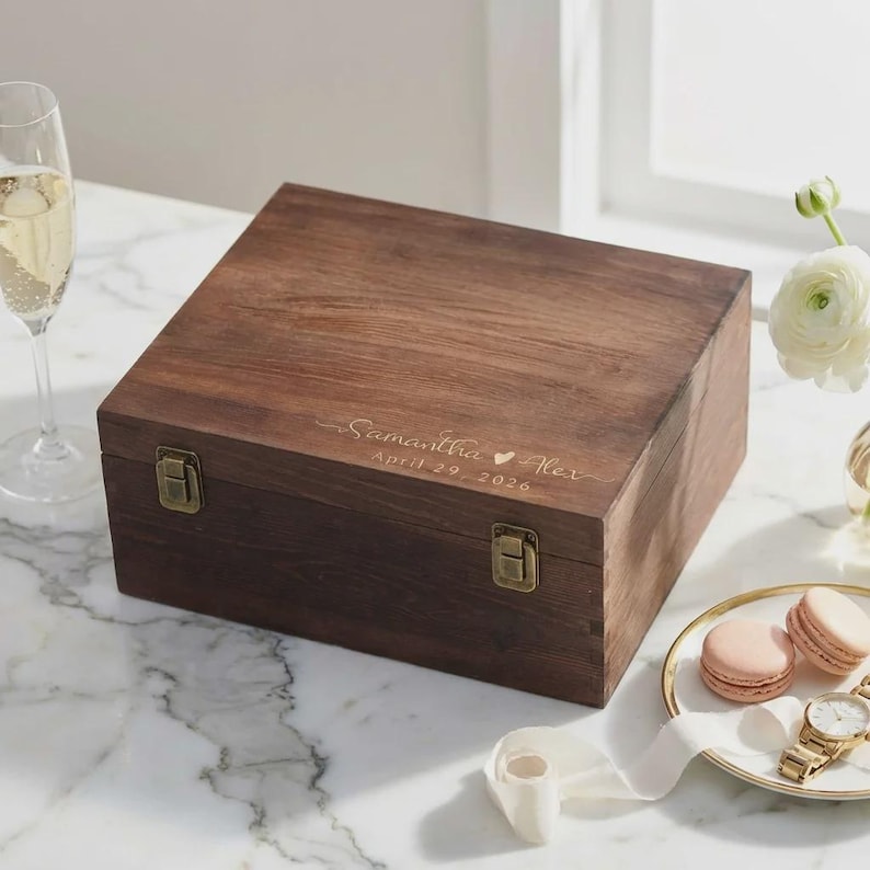 May include: A dark brown wooden box with a hinged lid and metal clasps. The box is engraved with the names "Samantha & Alex" and the date "April 29, 2026". The box is on a white marble surface.