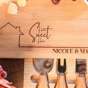 May include: A wooden cheese board with a house design, the words "Home Sweet Home" and the names "Nicole & Manhal" engraved on it. The board has four cheese knives with wooden handles.