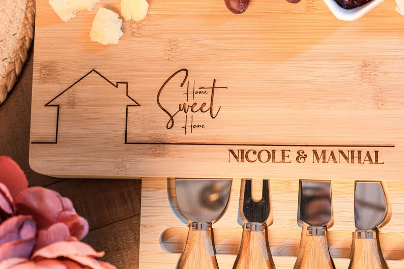 May include: A wooden cheese board with a house design, the words "Home Sweet Home" and the names "Nicole & Manhal" engraved on it. The board has four cheese knives with wooden handles.