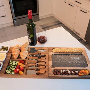 May include: A wooden serving board with a slate cheese board, four small bowls, and a variety of snacks, including crackers, nuts, dried fruit, and vegetables. The slate board has the text "Home Sweet Home" and "Tyler & Megan" engraved on it.