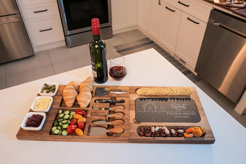 May include: A wooden serving board with a slate cheese board, four small bowls, and a variety of snacks, including crackers, nuts, dried fruit, and vegetables. The slate board has the text "Home Sweet Home" and "Tyler & Megan" engraved on it.