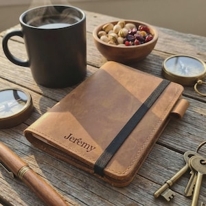 May include: A tan leather journal with the name "Jeremy" embossed on the cover, held closed by a black elastic band. A black mug with steam, a bowl of nuts, a compass, a pen, and keys are on a wooden surface.