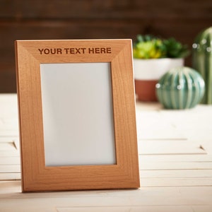 May include: A light brown wooden picture frame with the text "YOUR TEXT HERE" at the top. The frame is empty, with a white interior. Decorative ceramic cacti and succulents are in the background.