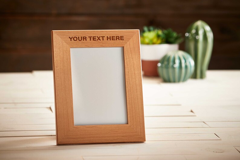 May include: A light brown wooden picture frame with the text "YOUR TEXT HERE" at the top. The frame is empty, with a white interior. Decorative ceramic cacti and succulents are in the background.