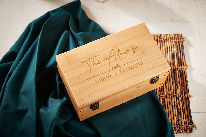 May include: A rectangular bamboo box with a hinged lid and metal clasp. The lid is engraved with "The Adams" above "Andrew + Samantha" and a floral design. The box rests on a dark teal fabric and a bamboo mat.
