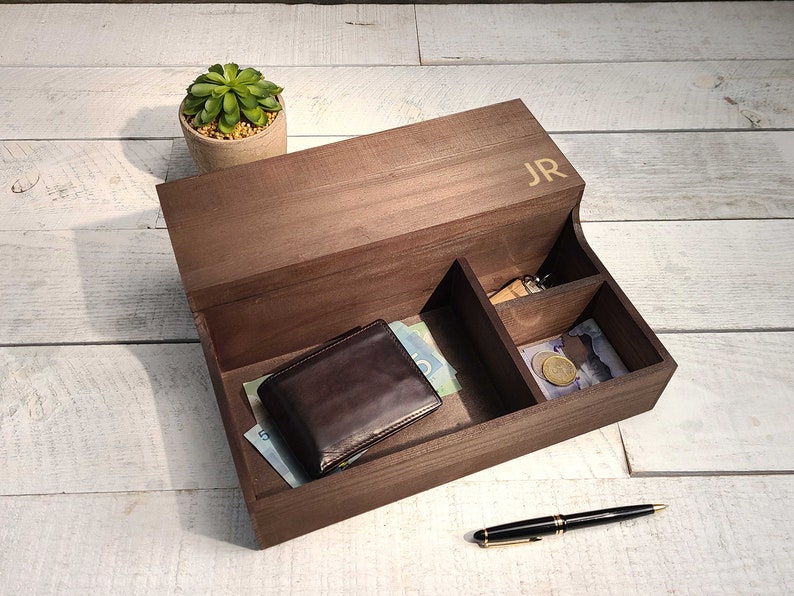 May include: A wooden box with compartments for organizing small items. The box has a lid with the initials "JR" engraved on it. The box is open and contains a wallet, cash, and a key.