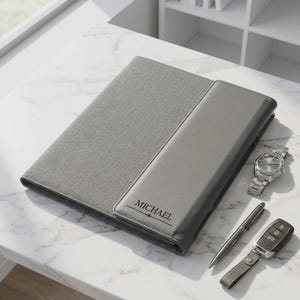 Peut inclure: Un folio gris avec le nom "MICHAEL" imprimé sur le devant, une montre-bracelet argentée, un stylo argenté et une clé de voiture avec une lanière en cuir sont présentés sur une surface en marbre blanc. Le folio a un tissu texturé et un matériau lisse semblable au cuir.