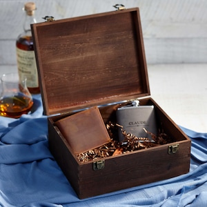 May include: A wooden gift box with a hinged lid, containing a brown leather wallet, a gray flask with the name "CLAUDE" engraved, and decorative filler. A bottle of liquor and a glass are in the background.