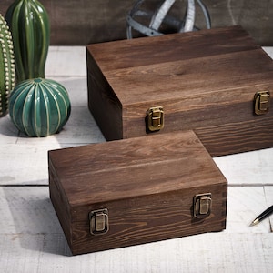 May include: Two wooden boxes with brass-colored clasps. The boxes are stained a dark brown and have a rectangular shape. A black pen with a gold-colored tip rests on the white-washed wooden surface. Decorative ceramic cacti are in the background.