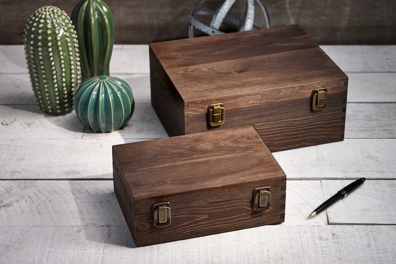 May include: Two wooden boxes with brass-colored clasps. The boxes are stained a dark brown and have a rectangular shape. A black pen with a gold-colored tip rests on the white-washed wooden surface. Decorative ceramic cacti are in the background.