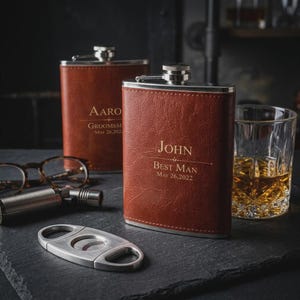 May include: Two brown leather hip flasks with silver tops, engraved with names and dates. A glass of amber liquid, spectacles, and a cigar cutter are also present. The flasks are suitable for a special event.