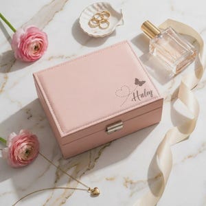 Peut inclure: Une boîte à bijoux rose pâle avec un motif de papillon et de cœur, le nom "Haley" est gravé sur le dessus. La boîte est fermée par un fermoir argenté. Des bagues dorées, un collier pendentif en forme de cœur et un flacon de parfum sont également visibles.