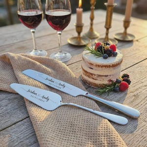 May include: A wedding cake with berries and rosemary sits next to a cake knife and server engraved with "Jordan & Andy 22/11/2025". Two glasses of red wine and candles are in the background. The cake knife and server are on a tan cloth.