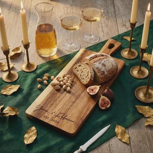 May include: A rectangular wooden serving board engraved with the text "Yuan & Rebecca EST. 2022" featuring sliced bread, pistachios, and halved figs. The board is placed on a dark green runner alongside a glass carafe, two wine glasses, and four brass candlesticks with cream-colored taper candles.