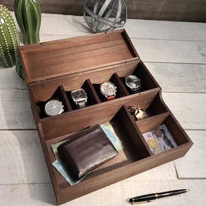 May include: A wooden box with a hinged lid, divided into compartments for storing watches, jewelry, or other small items. The box is open and shows five watches in the compartments, along with a wallet, keys, and coins.