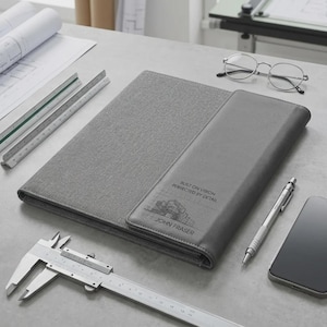 May include: A grey portfolio with the text "BUILT ON VISION PERFECTED BY DETAIL" and "JOHN FRASER" is open on a desk. Drafting tools, a pen, a smartphone, and spectacles are also on the desk.