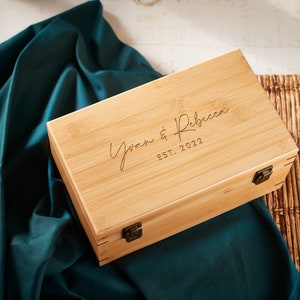 May include: A natural wood box with a hinged lid and brass clasps. The box is engraved with the text "Yvan & Rebecca EST. 2022".