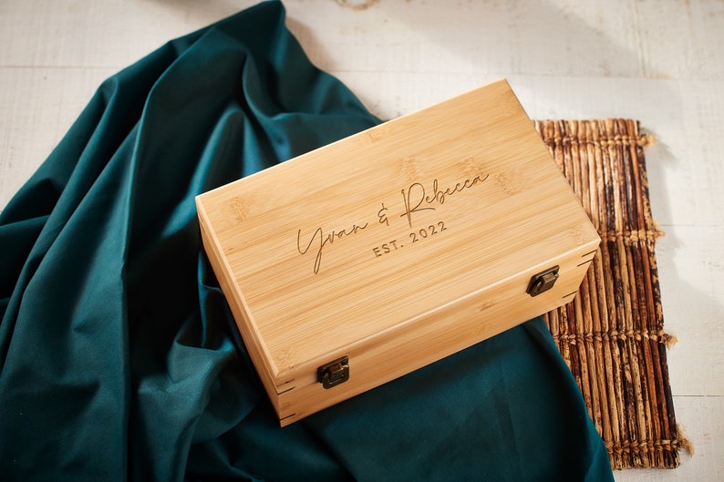 May include: A natural wood box with a hinged lid and brass clasps. The box is engraved with the text "Yvan & Rebecca EST. 2022".