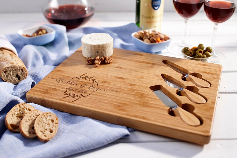 May include: A personalized bamboo cheese board with three built-in cheese knives. The board is engraved with the text "Heather & Bryan Aug 8, 2021" in a decorative font.