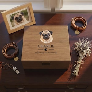 May include: A square wooden keepsake box featuring a printed illustration of a pug, the name CHARLIE, a paw print icon, and the text Always in our hearts. The box has a metal latch and a smooth wood grain finish, centred on a dark surface.