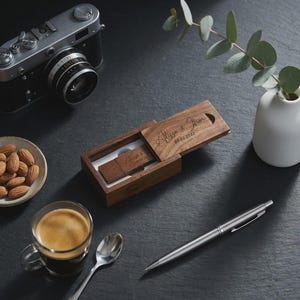 Peut inclure: Une clé USB et son étui en bois, gravés des noms "Alyssa & Jhon" et de la date "05.02.2022". L'étui est ouvert, révélant la clé USB à l'intérieur. Autres éléments : un appareil photo vintage, une tasse de café, des amandes, un stylo et un petit vase avec de la verdure.