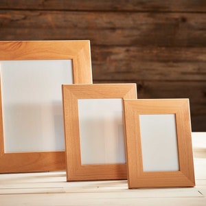 May include: Three wooden picture frames of varying sizes, with a light brown finish and white interiors, are displayed on a white surface. Decorative ceramic cacti and a round teal vase are in the background. The frames are ideal for displaying photos or artwork.