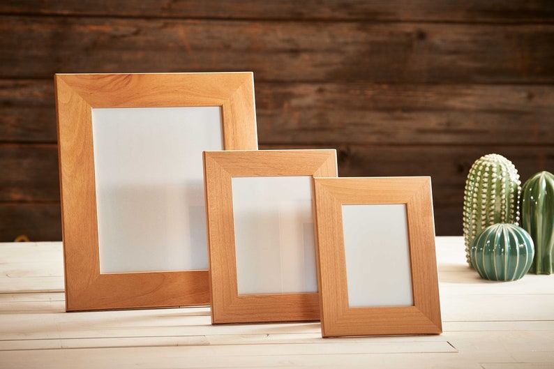 May include: Three wooden picture frames of varying sizes, with a light brown finish and white interiors, are displayed on a white surface. Decorative ceramic cacti and a round teal vase are in the background. The frames are ideal for displaying photos or artwork.