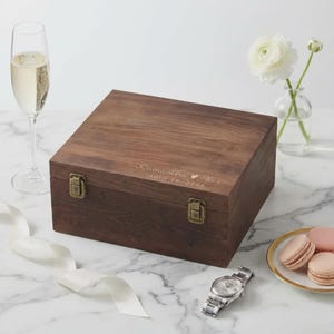 May include: A dark brown wooden box with metal clasps, engraved with the names "Samantha & Hex" and the date "April 29, 2026". A champagne flute, a watch, flowers, and macarons are also present.