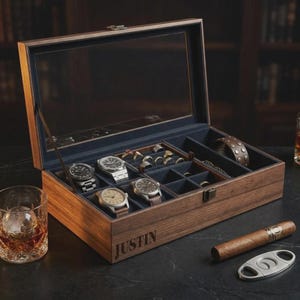 May include: A wooden watch and jewellery box with a glass lid, containing several watches, rings, and a bracelet. The box is open, revealing a blue interior. The name "JUSTIN" is engraved on the front. A glass of whisky, a cigar, and a cutter are on the table.
