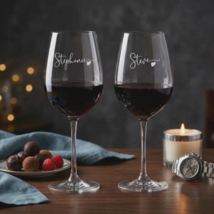 May include: Two wine glasses filled with red wine, etched with the names "Stephanie" and "Steve" in cursive, each with a heart. A lit candle, a watch, and a plate of chocolates and raspberries are on the table. The scene is set on a wooden surface.