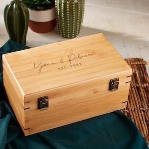 May include: A natural wood box with a hinged lid and brass clasps. The box is engraved with the text "Yvan & Rebecca EST. 2022".