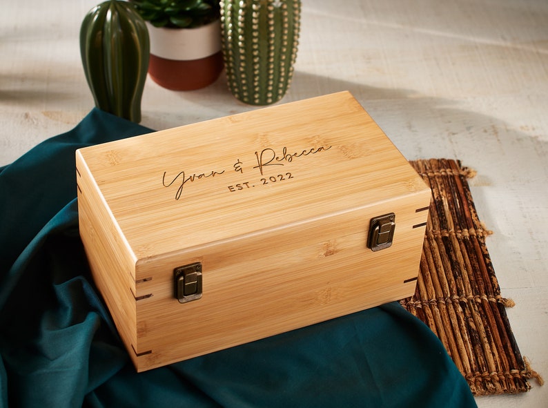 May include: A natural wood box with a hinged lid and brass clasps. The box is engraved with the text "Yvan & Rebecca EST. 2022".