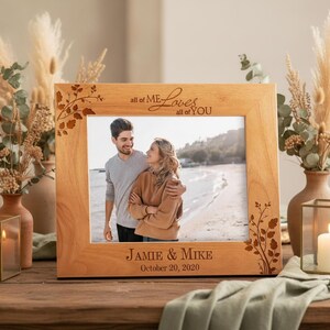 May include: A light-colored wooden picture frame with the text "all of ME Loves all of YOU" and "JAMIE & MIKE October 20, 2020" engraved on it. The frame has a floral design on the corners and holds a photo of a couple.