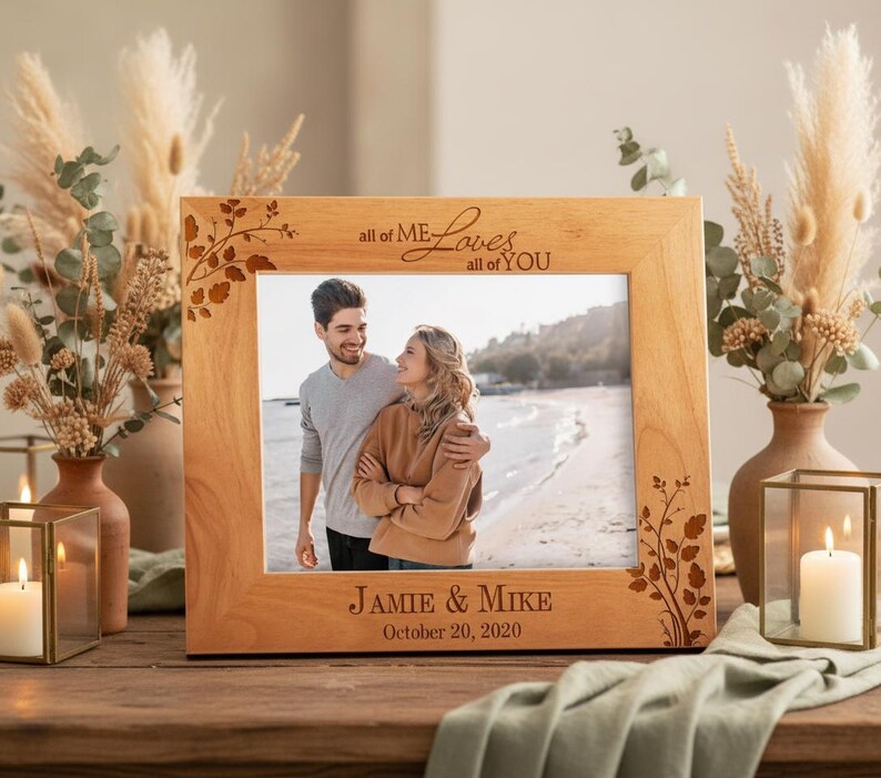 May include: A light-colored wooden picture frame with the text "all of ME Loves all of YOU" and "JAMIE & MIKE October 20, 2020" engraved on it. The frame has a floral design on the corners and holds a photo of a couple.