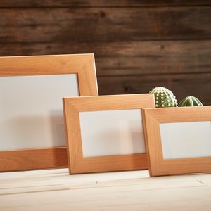 May include: Three wooden picture frames of varying sizes, arranged on a light-colored surface. The frames are a warm, natural wood tone, with a simple, rectangular design. The background is a dark wood panel. Small cacti are placed in the frames.