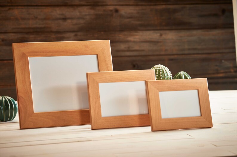 May include: Three wooden picture frames of varying sizes, arranged on a light-colored surface. The frames are a warm, natural wood tone, with a simple, rectangular design. The background is a dark wood panel. Small cacti are placed in the frames.